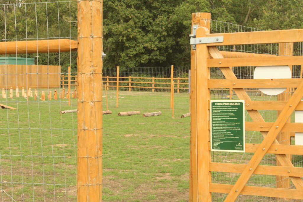 The gated entrance to the dog park at Rockingham Forest Park, with the dog park rules sign on the gate and carved timber posts and logs on the grass beyond