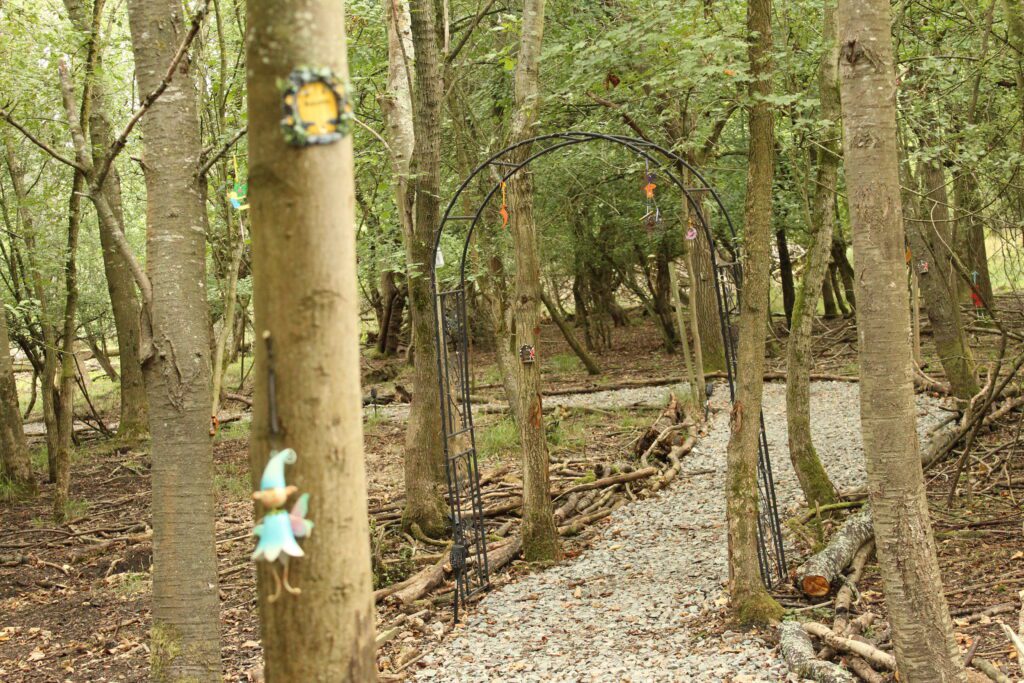 A fairy in a blue hat on a tree trunk beside the iron archway on the Fairy Fauna Trail, with wind chimes and small figures hanging from it