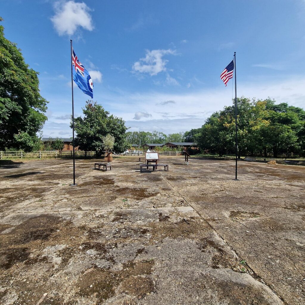 The concrete hangar base at Kings Cliffe, with the Union flag and the Stars and Stripes flying beside the Glenn Miller memorial.