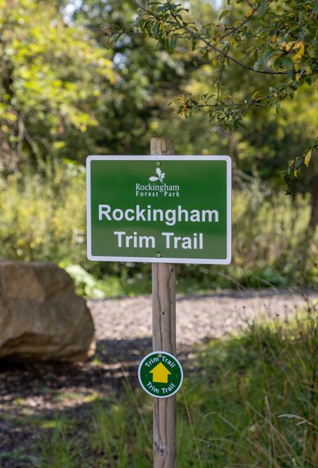 A signpost at the start of the trim trail: a green Rockingham Forest Park sign reading Rockingham Trim Trail, with a yellow waymarker disc on the post below it