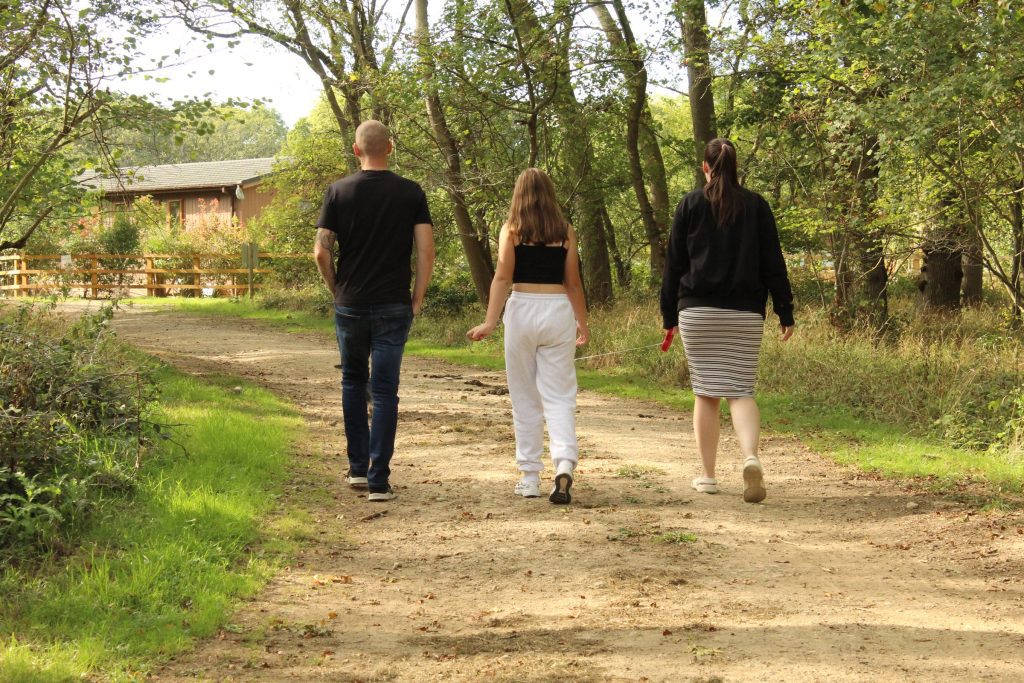 Three people walking away along a gravel path through the woods at Rockingham Forest Park, with a lodge among the trees behind them