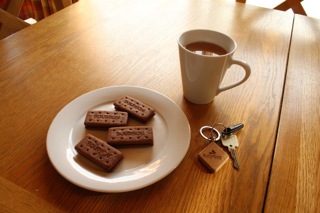 A mug of tea, a plate of biscuits and the lodge keys on the dining table inside a lodge at Rockingham Forest Park