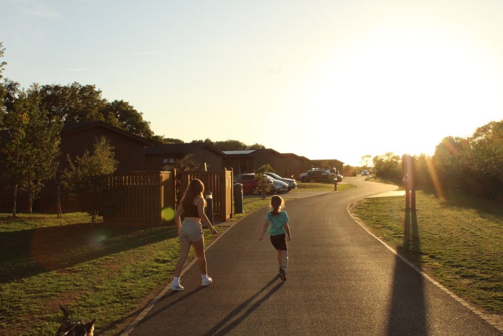 Two guests walking down the lodge road at Rockingham Forest Park in low evening sun, lodges on one side and open grass on the other
