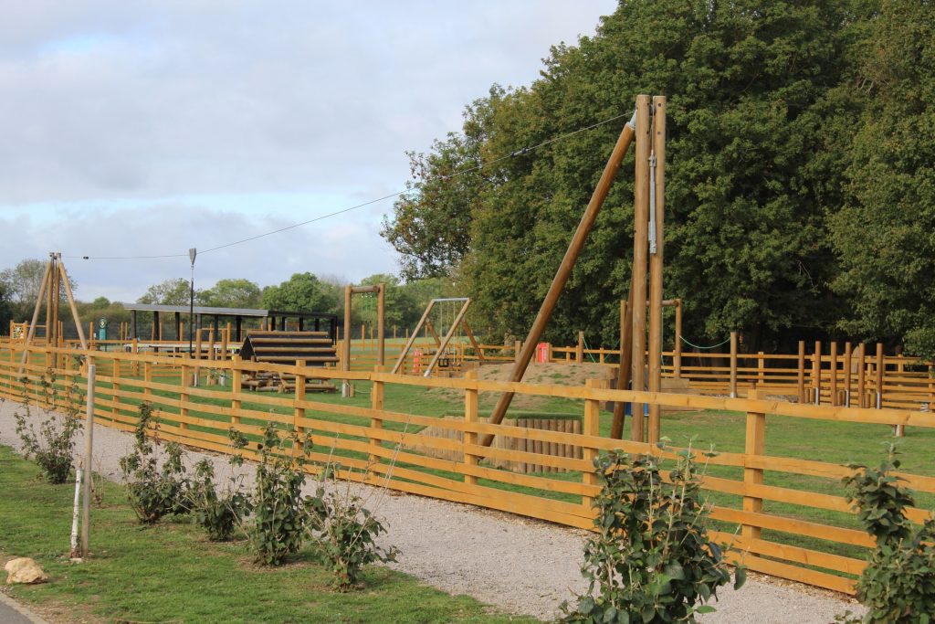 The play park at Rockingham Forest Park, seen from the path: timber climbing frames and swings inside a low post-and-rail fence, with the shelter behind